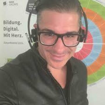 Mann mit Brille und Headset lächelt in die Kamera, hinter ihm ein Plakat der WBS GRUPPE.