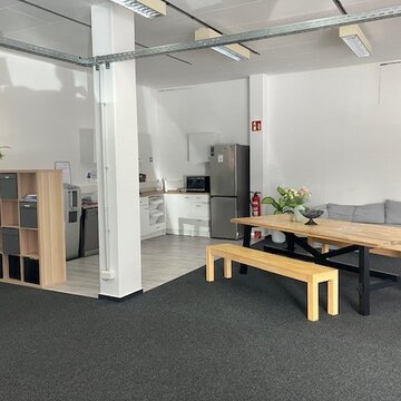 Küche mit Kühlschrank, Wasserspender, Regal mit Fachablage und Tisch mit zwei Sitzbänken sowie ein graues Sofa.
