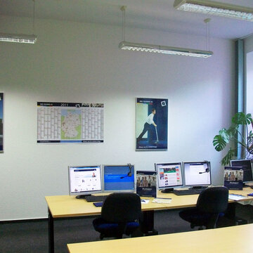 Seminarraum mit 3 Arbeitsplätzen jeweils mit 2 Bildschirmen, einem Bürostuhl, Tastatur, Maus und Headset. An der Wand Plakate und ein Wandkalender.