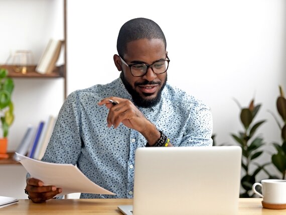 Ein Mann sitzt vor seinem Laptop und hält Papier und einen Kugelschreiber in den Händen.