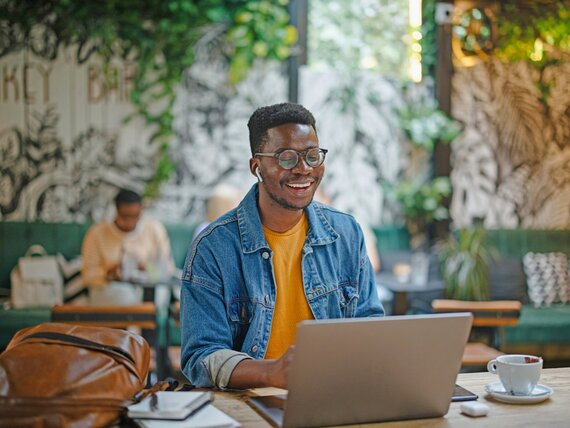 Ein fröhlich lachender Mann mit Kopfhörern sitzt in einem Café vor seinem Laptop.