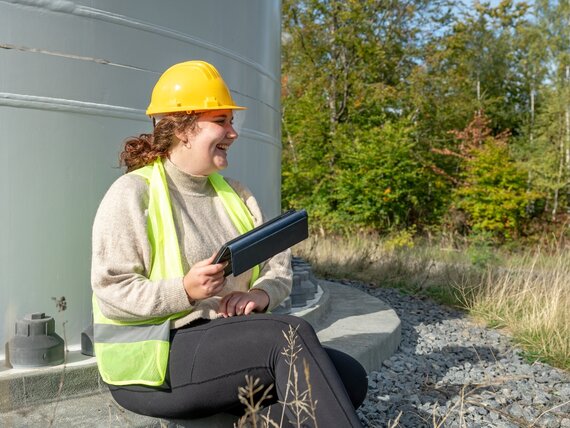 Eine fröhliche Frau, die einen Helm und eine Schutzweste trägt sitzt draußen mit ihrem Tablet.