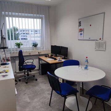 Prüfungsraum mit rundem Tisch und 3 Stühlen. Vor dem Fenster befindet sich ein zusätzlicher Arbeitsplatz mit PC.