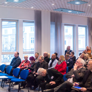 Eine Frau hält einen Vortrag und steht an einem Rednerpult. Vor ihr sitzen ca. 50 bis 80 Personen, die dem Vortrag folgen.