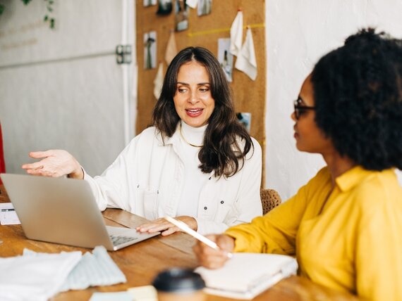 Eine Frau erklärt einer anderen Frau etwas am Laptop während diese sich Notizen macht.