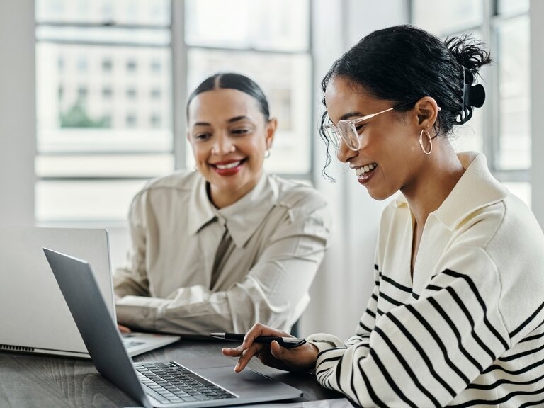 Zwei Frauen arbeiten fröhlich an ihren Laptops.