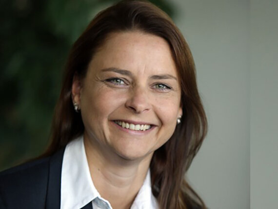 Eine brünette Frau mit langen Haaren. Sie trägt eine weiße Bluse und einen dunklen Blazer.
