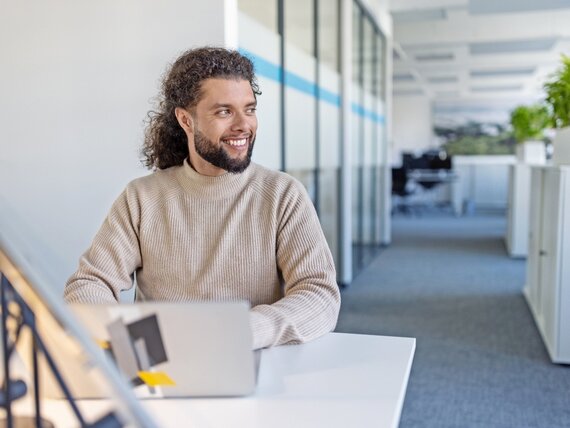 Ein fröhlicher Mann mit langen Haaren und Bart sitzt in einem Großraumbüro vor seinen Laptop