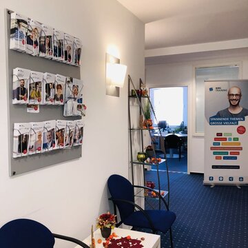 Gang mit zwei Stühlen und einem kleinen Tisch. Dahinter an der Wand befindet sich eine Halterung mit vielen Flyern mit Informationsamterialien.