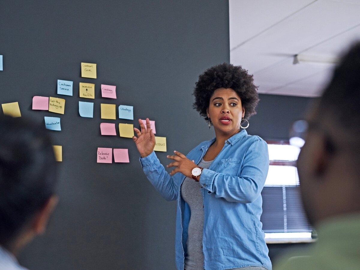 Eine Frau steht vor einer Wand mit bunten Notizzetteln und hält einen Vortrag.