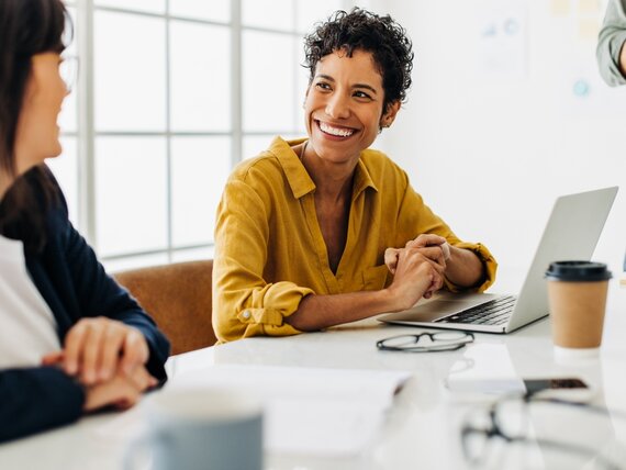 Zwei Frauen sitzen mit ihren Laptop und Notizbuch gut gelaunt in einem Meeting