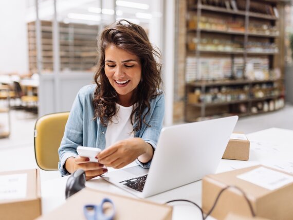 Eine Frau sitzt in einem Geschäft und arbeitet am Laptop mit ihrem Smartphone in der Hand.