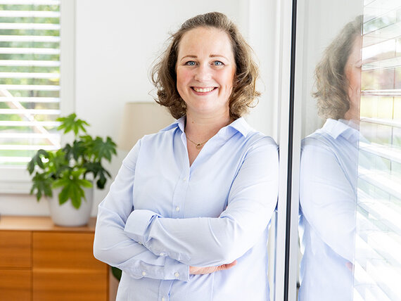 Coachin Verena Müller Eine Frau mit heller Bluse steht mit verschränkten Armen vor einem Fenster. Ihr Gesichtsausdruck ist freundlich, hinter ihr steht eine Topfpflanze.