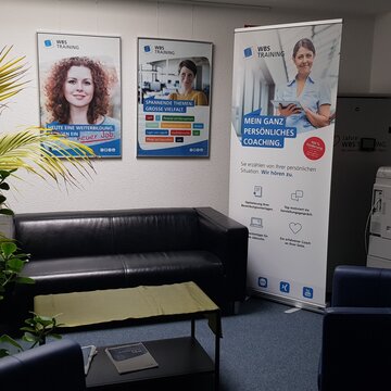 Aufenthaltsbereich mit Ledercouch und Sesseln, einer großen, grünen Zimmerpflanze sowie Druckstation. Zusätzlich steht ein Werbeaufsteller im Bereich.