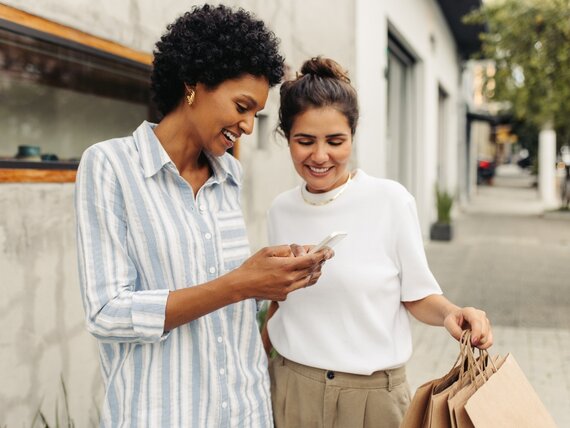 2 Frauen mit Einkaufstaschen stehen draußen und schauen sich etwas auf dem Smartphone an.