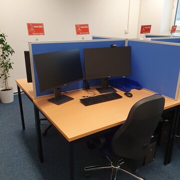 Ein Schulungsraum mit mehreren Arbeitsbereichen, die durch blaue Trennwände voneinander getrennt sind. Jeder Arbeitsplatz besteht aus 2 PC-Bildschirmen, Tastatur und Maus sowie Headset.