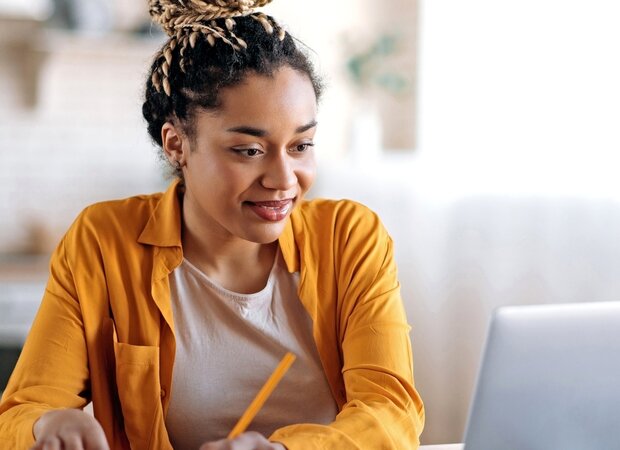Eine Frau sitzt in der Küche vor ihrem Laptop und macht sich Notizen.