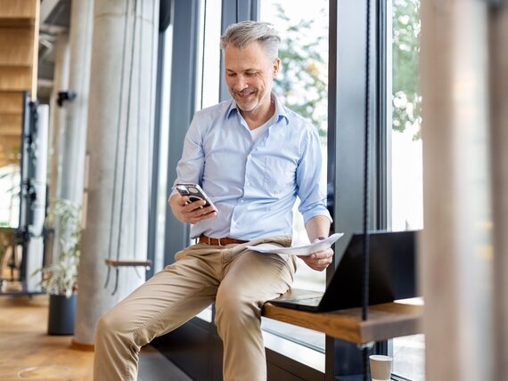 Ein Manager sitzt mit seinem Laptop und Unterlagen an einem Fenster und schaut auf sein Smartphone