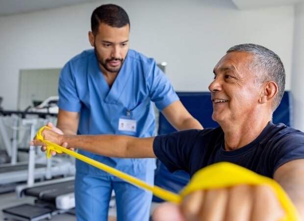 Ein Physiotherapeut unterstützt seinen Patienten bei Übungen mit einem gelben Gymnastikband