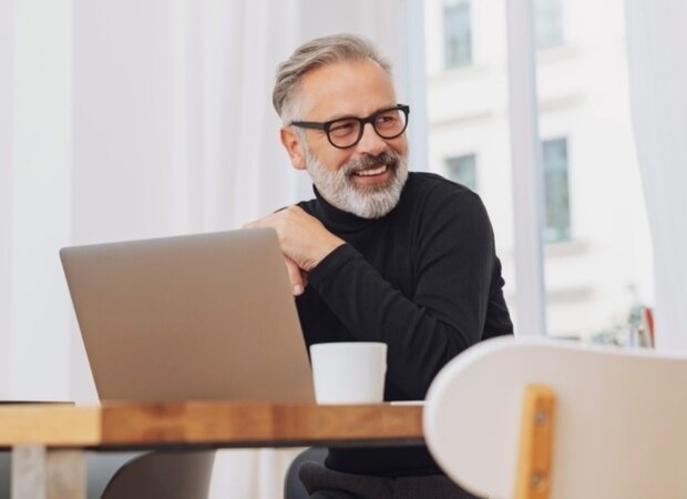 Ein fröhlicher Mann sitzt vor seinem Laptop und trinkt einen Kaffee.