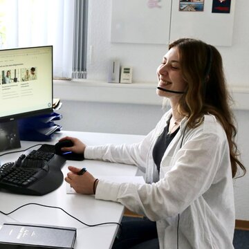 Eine Teilnehmerin sitzt an ihrem Schreibtisch vor dem PC und trägt ihr Headset während sie fröhlich lernt und sich Notizen macht.