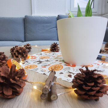 dekorierter Beistelltisch mit Lichterkette, Tannenzapfen und einer herbstlichen Servierte unter einem Blumentopf. Im Hintergrund steht ein graues Sofa.