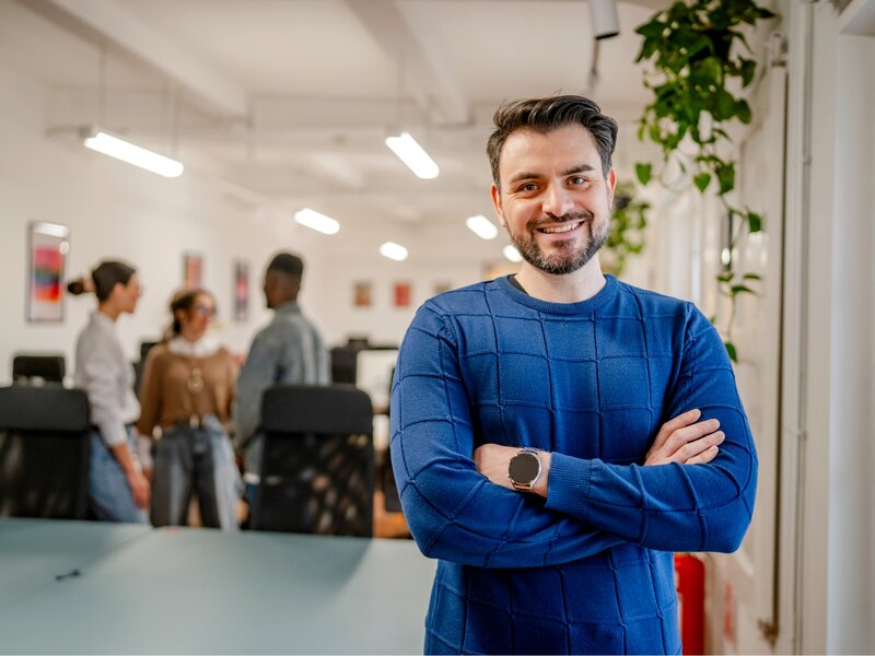 Mann in blauem Pullover steht mit verschränkten Armen in einem Lernraum. Weitere Personen im Hintergrund.