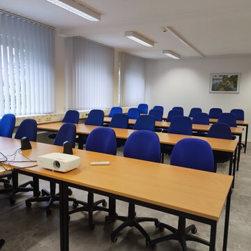Schulungsraum für den Präsenzunterricht mit Tischreihen und blauen Bürostühlen. Vorne steht ein Beamer auf dem Tisch.