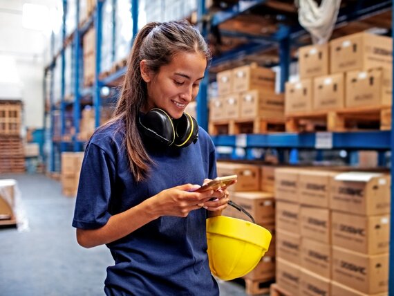 Eine Frau mit Hörschutz und Helm steht in einem Lager und tippt auf ihrem Smartphone.