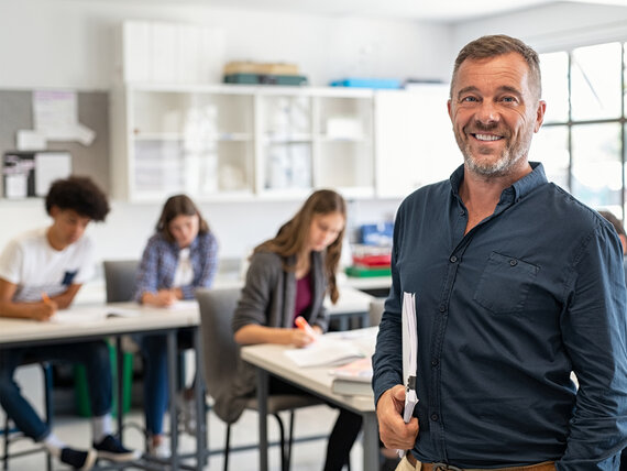 Lehrer steht in einem Unterrichtsraum vor einer Klasse mit einer Arbeitsmappe unter dem Arm.