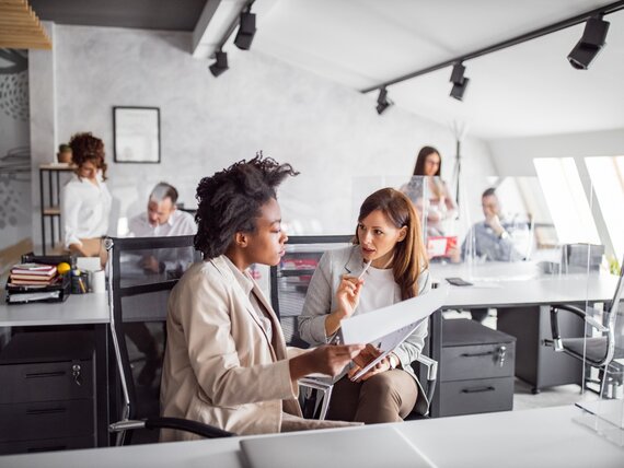 Zwei Frauen unterhalten sich in einem Großraumbüro über Dokumente