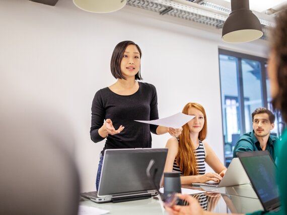 Eine junge Frau steht an einem Konferenztisch und erklärt ihrem Team etwas