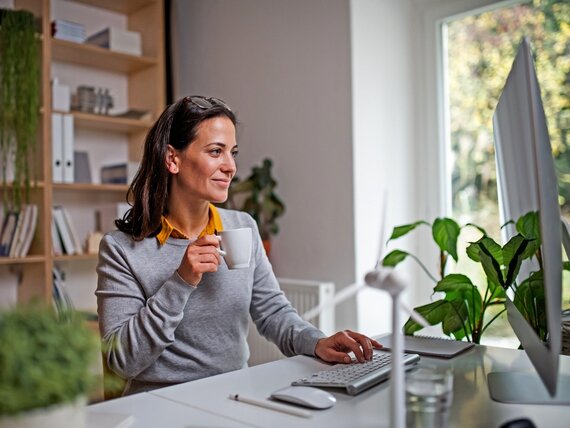 Eine Frau sitzt vor ihrem Computer und hält eine Kaffeetasse in der Hand.