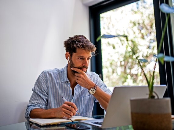 Ein Mann mit Kopfhörern in den Ohren sitzt vor seinem Laptop und macht sich Notizen.