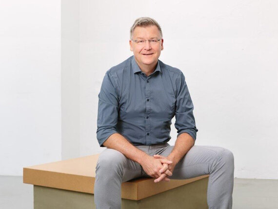 Aufnahme von Jens Grosse, der ein blaues Hemd und eine graue Jeans trägt und auf einem Hocker sitzt. 