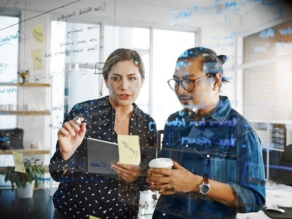 Eine Frau und ein Mann stehen vor einer Glaswand und sprechen über Notizen.
