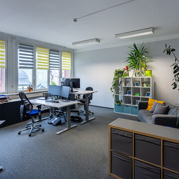 Mitarbeiterbüro mit mindestens 2 Arbeitsplätzen, mehreren Regalen im Raum. Rechts steht ein graues Sofa und mehrere Pflanzen schmücken den Raum.
