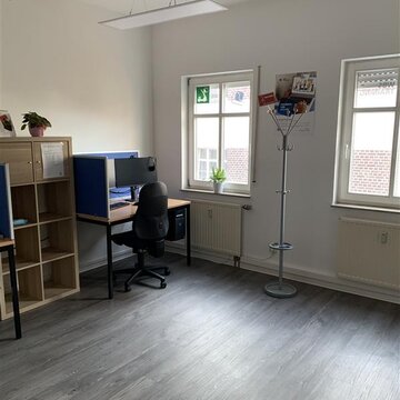 Raum mit rechts 2 Fenstern, zwischen denen ein Kleiderständer steht. Links ein Arbeitsplatz mit Bürostuhl, 2 Bildschirmen und Tastatur. links daneben ein hohes Holzregal mit Zimmerpflanze.