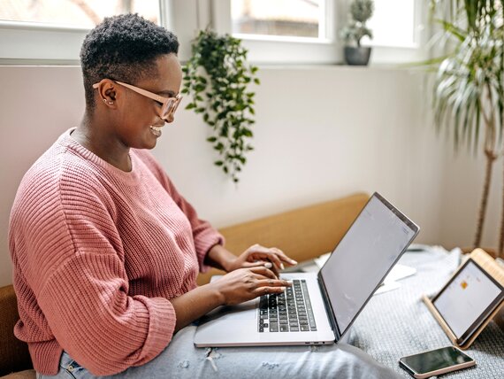 Eine Frau sitzt auf ihrem Bett und arbeitet am Laptop. 