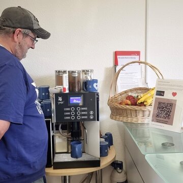 Ein Mann steht vor einem Kaffeevollautomat und macht sich einen Kaffee. Daneben ein Obstkorb sowie ein Aufsteller mit einem QR Code und ein kleiner Ventilator im Hintergrund.