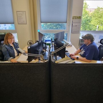 Empfangsbereich mit 2 Mitarbeitenden, die an ihren Schreibtischen vorm Computerbildschirm sitzen und gut gelaunt arbeiten. Geöffnetes Fenster mit Blick ins Grüne.