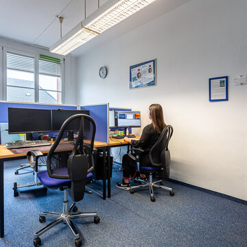 Raum mit mehreren Arbeitsplätzen mit jeweils 2 Bildschirmen, einem Bürostuhl, Tastatur und Maus. Am rechten Platz sitzt eine Frau und arbeitet. An der Wand hängen Plakate und eine Uhr. Der Boden ist mit blauem Teppich ausgelegt.