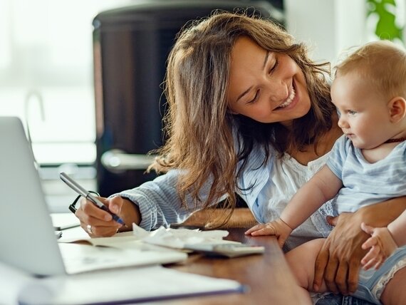 Eine Frau im Homeoffice mit ihrem Baby auf dem Arm sitzt vor ihrem Laptop.