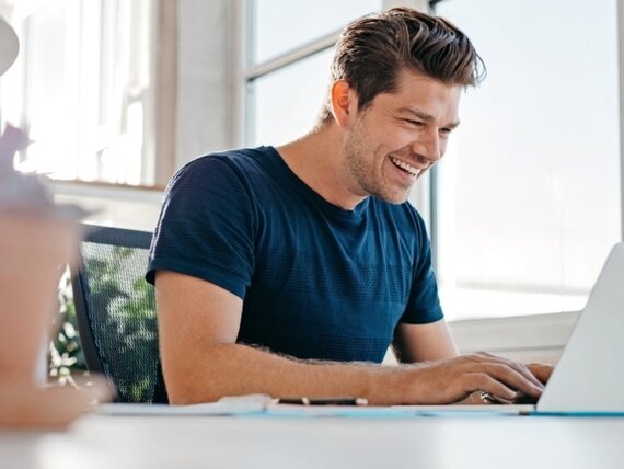 Ein fröhlicher Mann im Homeoffice bei der Arbeit an seinem Laptop.
