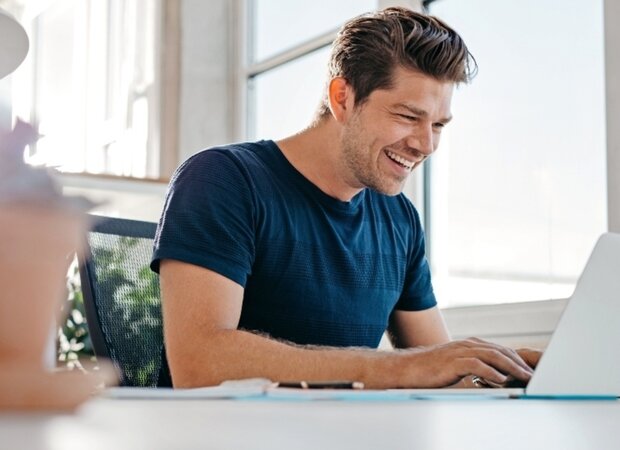 Ein fröhlicher Mann im Homeoffice bei der Arbeit an seinem Laptop.