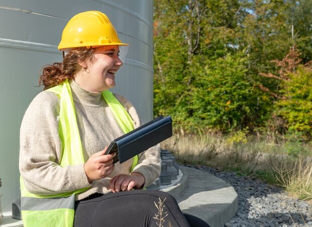Eine fröhliche Frau, die einen Helm und eine Schutzweste trägt sitzt draußen mit ihrem Tablet.