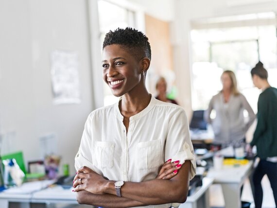 Eine lächelnde Frau steht in einem offenen Büro mit anderen Personen im Hintergrund. 