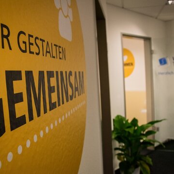Eingangsbereich. Glastüre mit WBS TRAINING Aufschrift und Logo. Zimmerpflanze. Gelbes Wandtattoo mit der Aufschrift „Wir gestalten gemeinsam“.