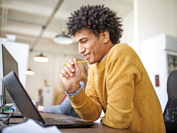 Ein fröhlicher Mann schaut auf seinen Laptop und hält einen Stift in der Hand.