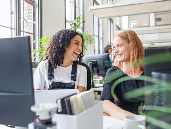 Zwei Frauen sitzen im Büro an ihren Computern und lachen fröhlich.